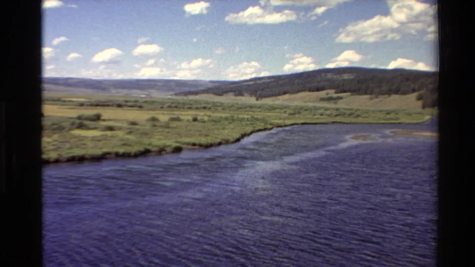 1980 8mm rural Western U.S. view showing a curving water edge, open grassland, and a dark wooded slope beyond.
