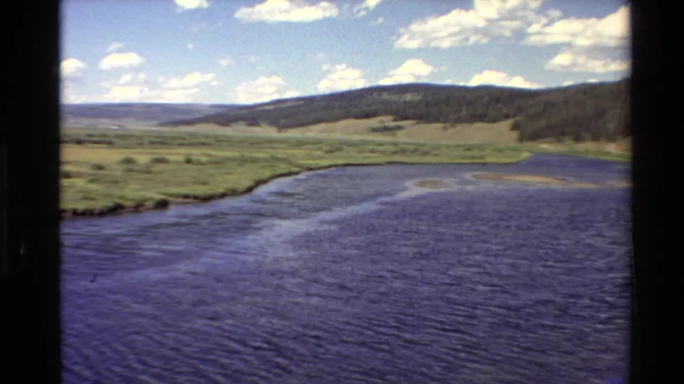Western U.S. home-movie frame from 1980 with rippled water, green shoreline, rolling hill, and scattered white clouds.