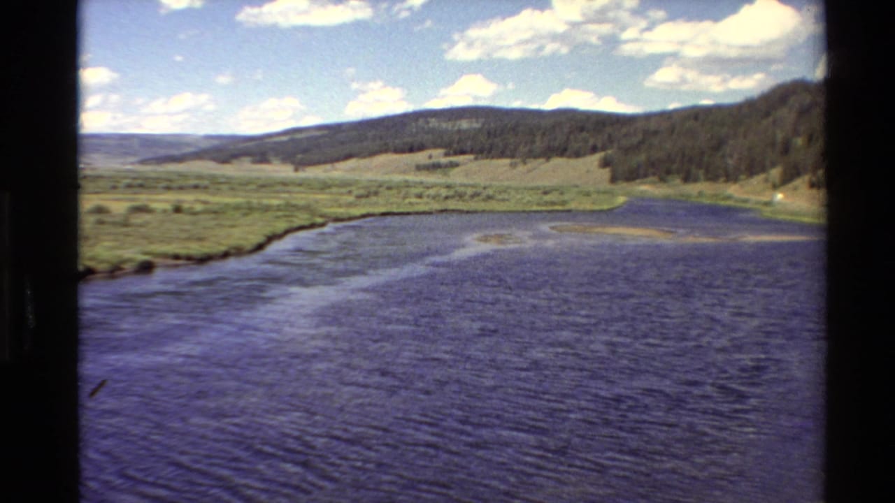 1980 Western U.S. 8mm frame showing a broad river, grassy valley edge, forested hillside, distant ridge, and bright clouded sky.