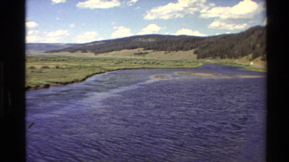 1980 Western U.S. 8mm frame showing a broad river, grassy valley edge, forested hillside, distant ridge, and bright clouded sky.