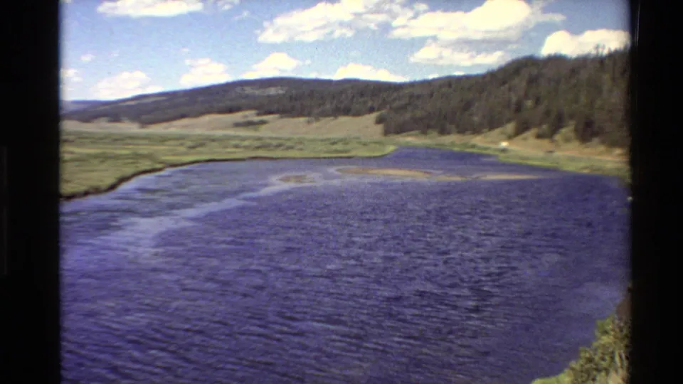 Wide 8mm landscape view of a Western U.S. river valley in 1980, with rippled water, meadow, wooded slope, distant ridge, and scattered clouds.