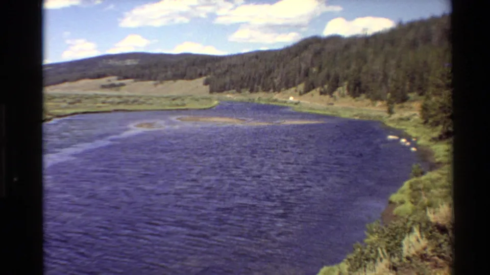 The pan reveals more of the curving river and right bank, with pine trees, grassy slope, open meadow, and bright sky in the 1980 home-movie frame.