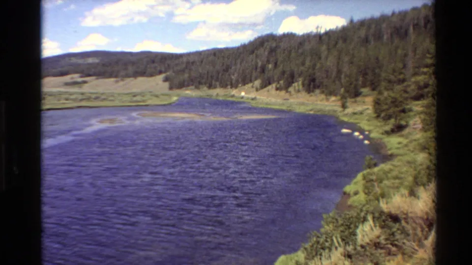 Late-pan 8mm frame from the 1980 Western U.S. clip showing the river bend, near grassy bank, pine-covered hillside, meadow, and clouded blue sky.