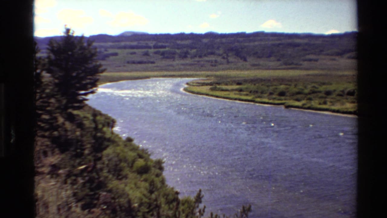 1980 Western USA home-movie frame of a broad river bend, pine-lined bank, green valley, and distant hills under bright sky.