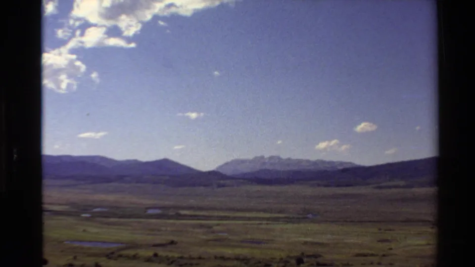 1980 Western USA home-movie frame showing open plains, small water patches, distant mountains, and scattered clouds.
