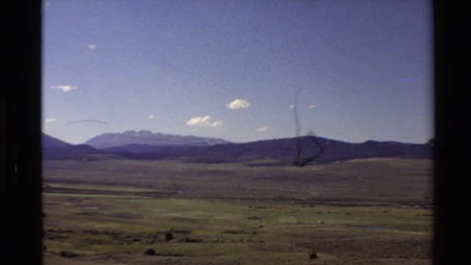 Western U.S. home-movie view of open grassland, low dark hills, distant mountains, blue sky, and visible scratches in the film image.