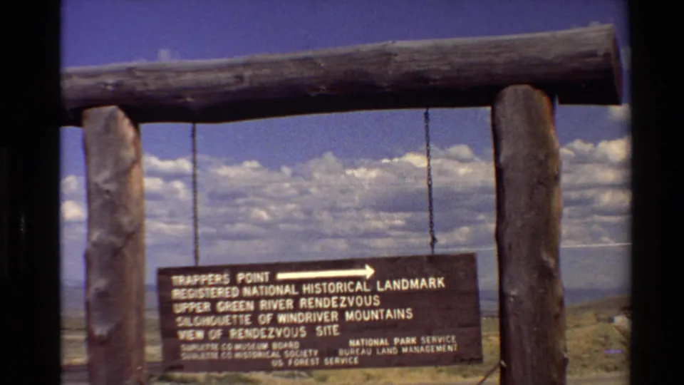 8mm frame of a rustic Trappers Point sign suspended from log supports, with bright clouds and distant terrain beyond the overlook.