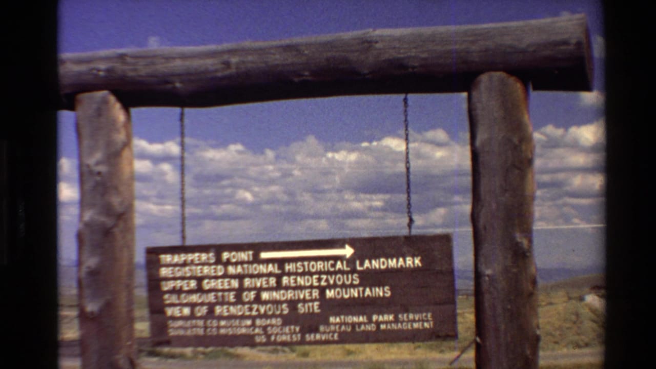 1980 8mm frame of a Trappers Point landmark sign between log posts with clouds and distant Western U.S. terrain behind it.