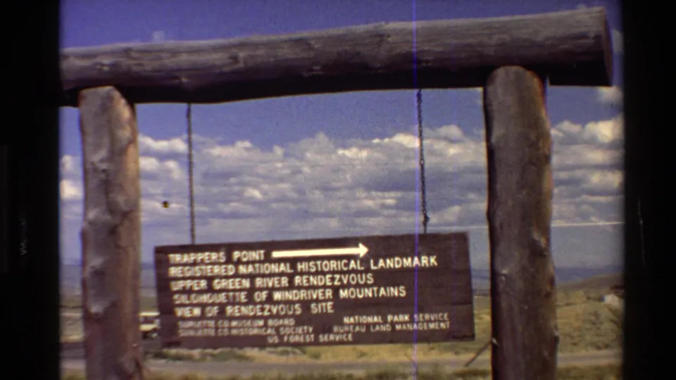 1980 8mm frame with the hanging Trappers Point sign partly lower in view, framed by logs and a bright clouded sky.