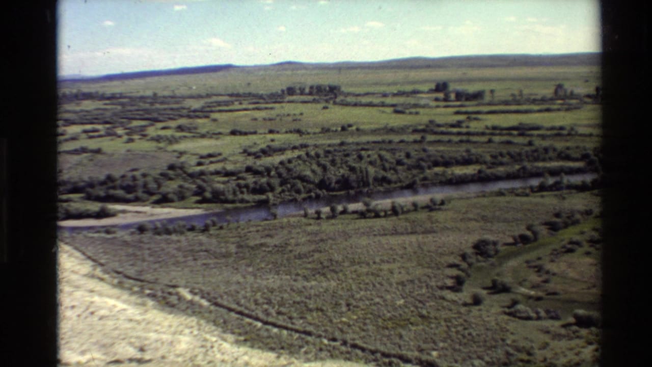 1980 Western U.S. 8mm aerial view over a winding river, open pasture, tree lines, and low hills under a bright sky.