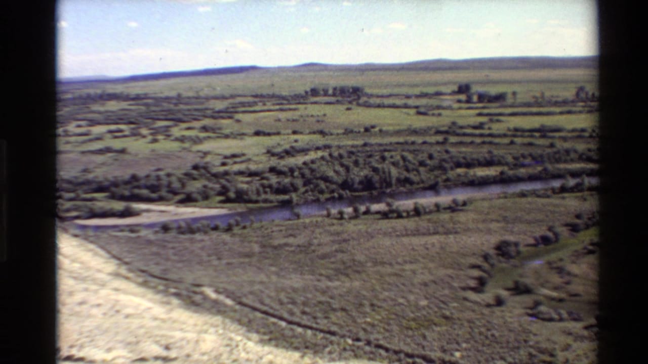 Aerial river valley with open pasture, trees, buildings, and low hills in the distance.