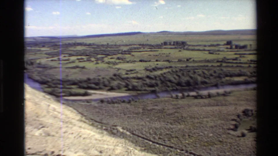 1980 8mm aerial frame over a pale foreground slope, river bend, green-brown fields, tree line, and low horizon hills.