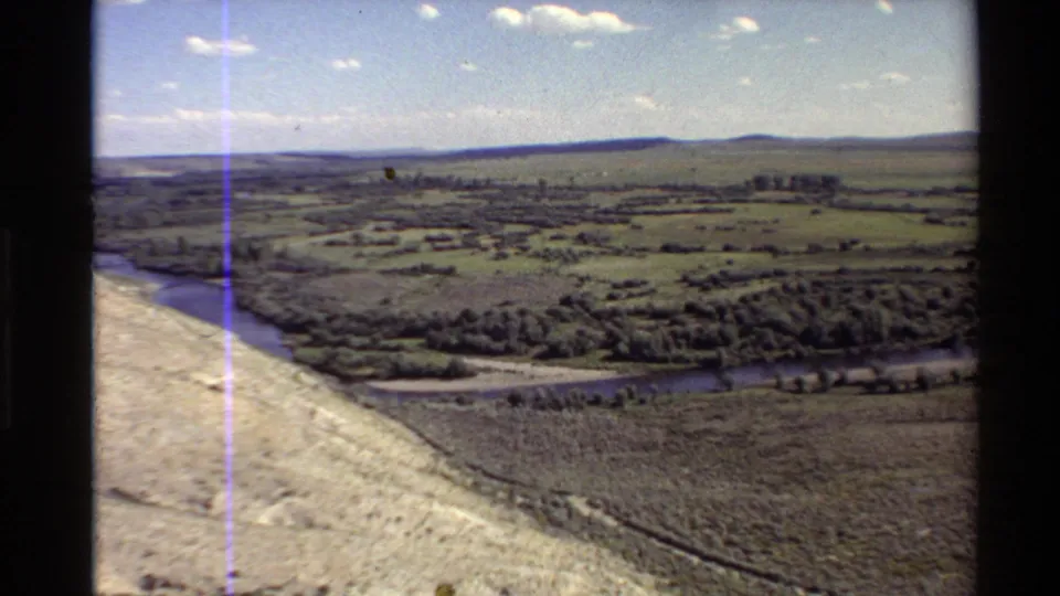 Wide 8mm aerial view of a Western U.S. river valley with pasture, tree rows, distant buildings, low hills, and film scan artifacts.