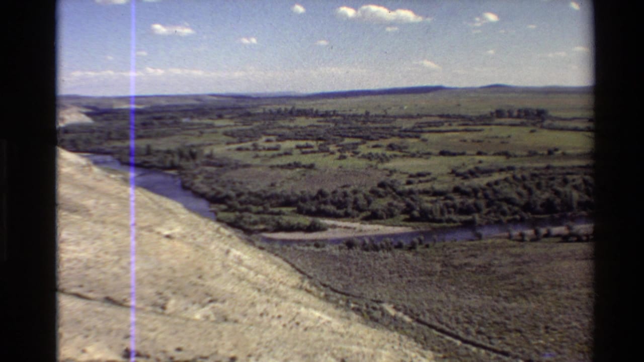 1980 8mm aerial view in the Western United States showing a river valley, pale hillside, fields, trees, and distant ridges.