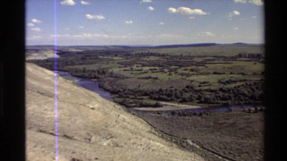 Aerial 8mm frame of Western U.S. countryside with a river, hillside, fields, tree belts, and a bright clouded sky.