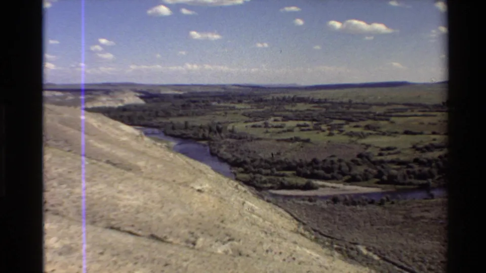 1980 home-movie aerial footage shows a Western U.S. river valley bordered by pale slopes, green fields, and tree lines.