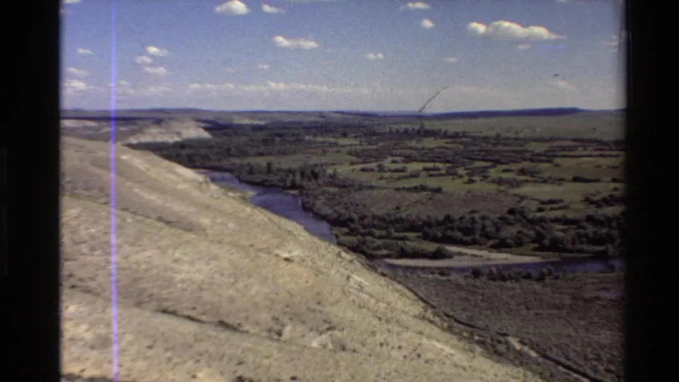 Western U.S. 8mm aerial frame with river, hillside, fields, trees, distant ridges, clouds, and a visible purple scratch line.