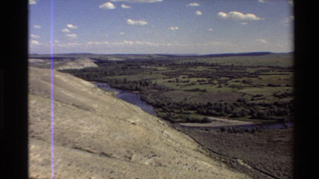 1980 8mm aerial view in the Western U.S. showing a pale slope, winding waterway, open fields, scattered trees, and a clouded sky.