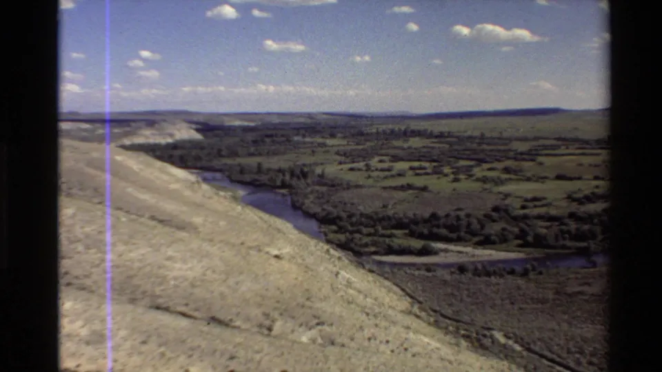 1980 8mm aerial view in the Western U.S. showing a pale slope, winding waterway, open fields, scattered trees, and a clouded sky.