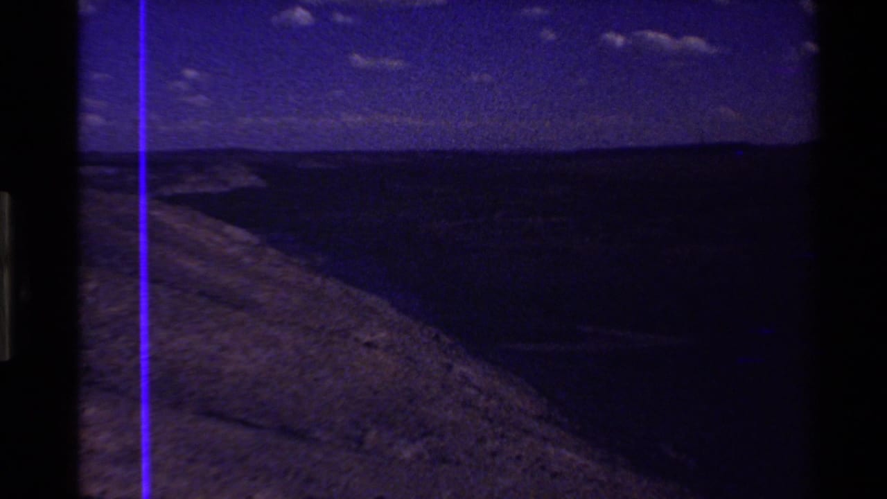 Blue-toned 8mm aerial frame of a hillside and distant ridge in the Western U.S., with heavy underexposure and a vertical purple film line.