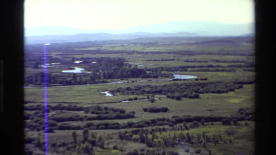 1980 8mm aerial view in the Western U.S. showing green plains, small ponds, scattered tree clusters, distant mountains, and faded color.