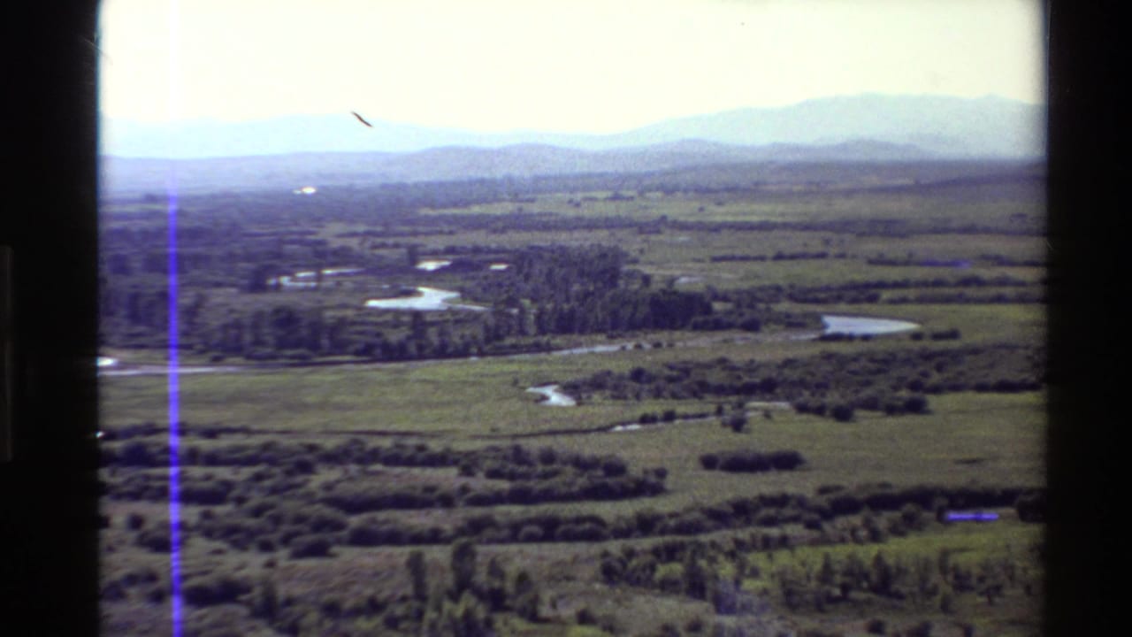 1980 Western USA 8mm aerial view of a winding river through grassland, ponds, shrubs, tree lines, and hazy mountains.