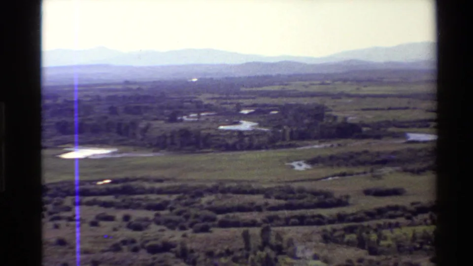 Aerial 8mm view over Western USA grassland, with a winding waterway, bright pond, shrub patches, trees, and distant ridges.