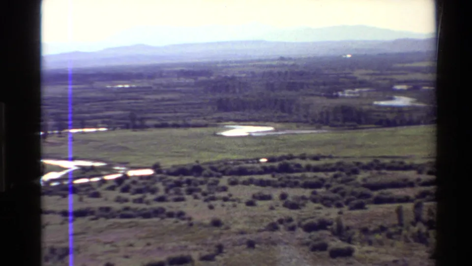 1980 8mm aerial frame showing bright river bends, wetland patches, open grassland, shrubs, tree rows, and hazy Western USA hills.