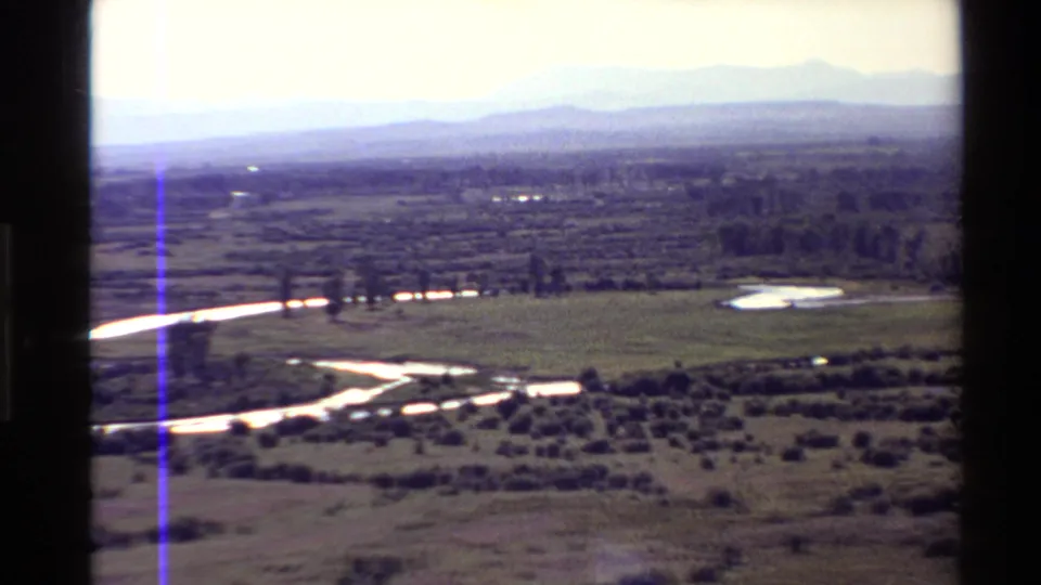 Western USA aerial home-movie frame with a bright winding river, pond, open grassland, shrubland, tree lines, and distant mountains.