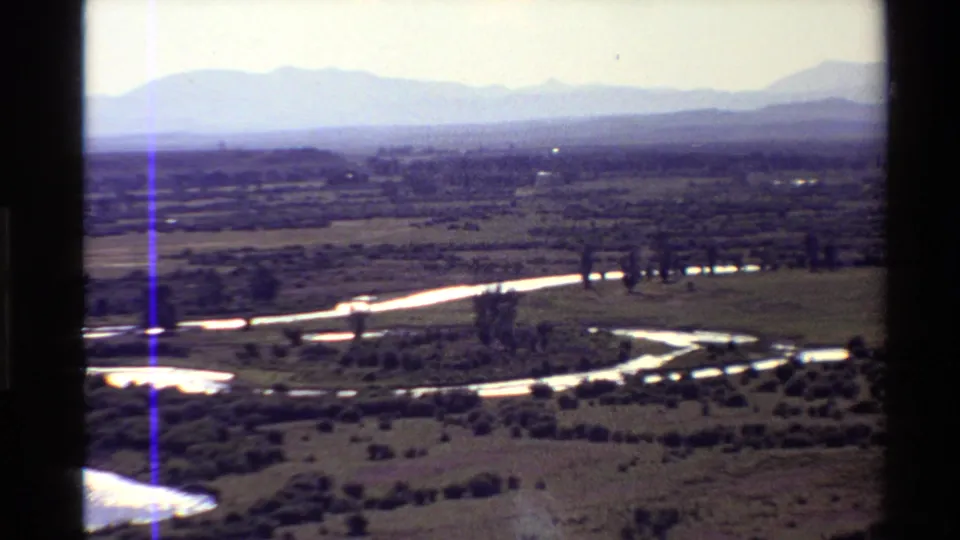 Vintage 8mm view of a meandering river bordered by trees and open Western U.S. plains with hazy mountains beyond.