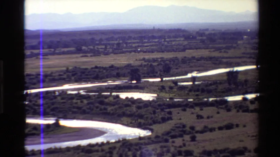 1980s 8mm frame showing a broad river curve, scattered vegetation, flat rural land, and distant Western U.S. mountains.