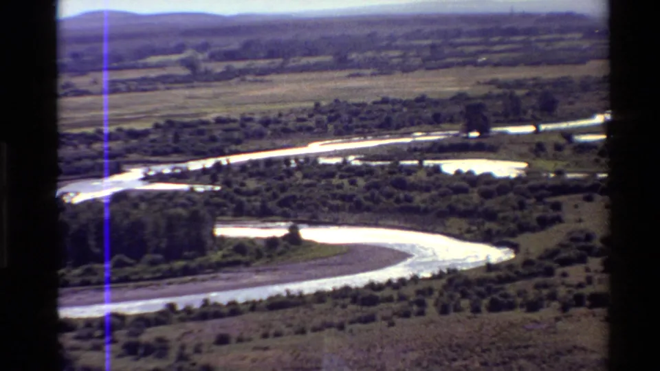 Elevated 8mm home-movie view of a curving river, tree-dotted flat land, and a hazy Western U.S. mountain backdrop.