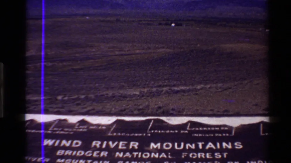 1980 8mm frame of a Western U.S. plain and distant ridge, with partial Wind River Mountains Bridger National Forest sign visible in front.