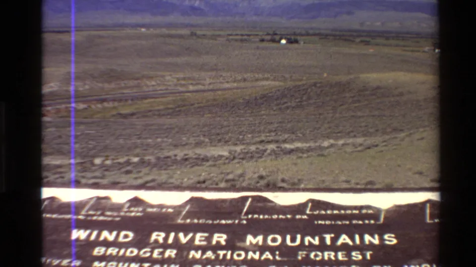 1980 home-movie frame showing a broad Western U.S. plain with distant mountains and partial Bridger National Forest sign lettering at the bottom.
