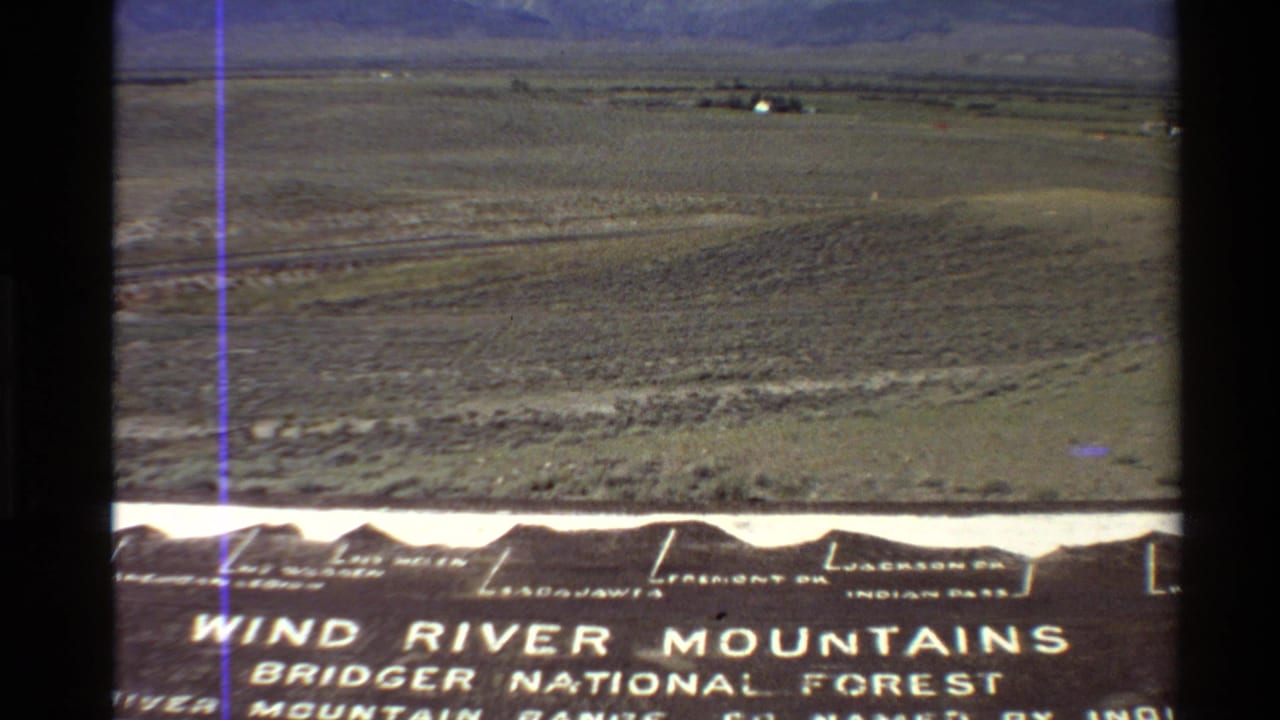 Roadside sign for Wind River Mountains and Bridger National Forest below a distant valley view.