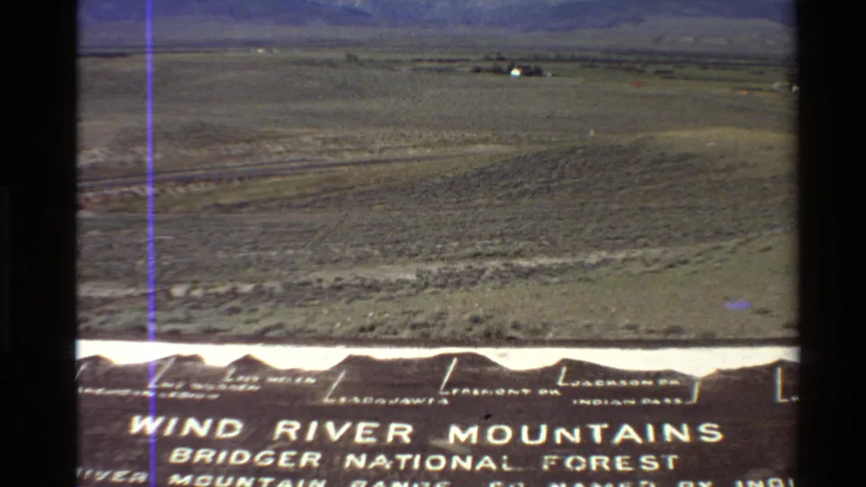 1980 8mm Western U.S. frame showing a roadside Wind River Mountains and Bridger National Forest sign with arid valley beyond.