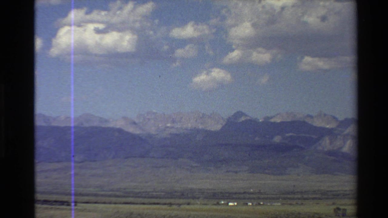 1980 Western USA home-movie frame showing desert plain and distant mountains beneath a partly cloudy sky, viewed through a dark window edge.