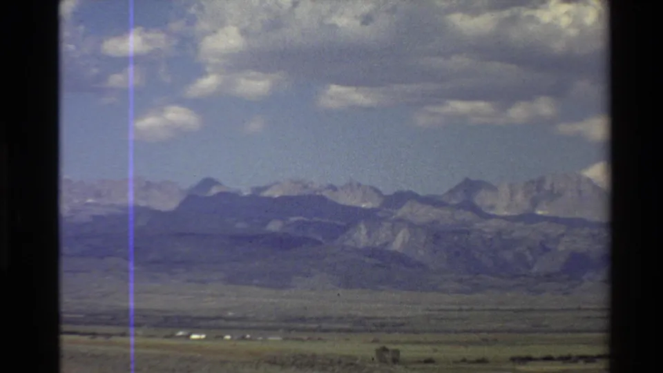 Home-movie view of a Western desert valley and distant mountain range, with cloudy sky, visible grain, and a purple vertical artifact.