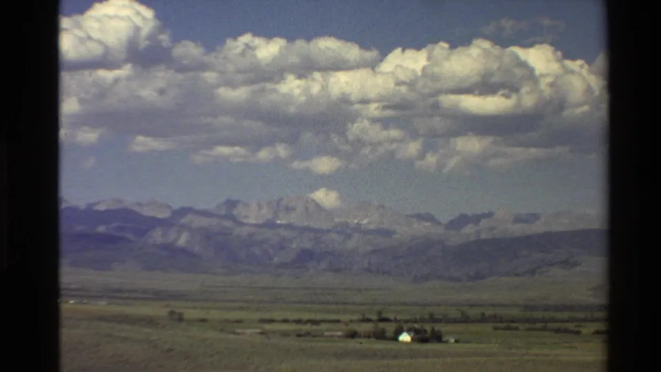 1980 home-movie frame showing a rural Western USA plain, small white structure, distant mountains, and broad cloud cover.