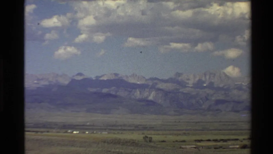 Vintage 8mm Western USA landscape view with a broad plain, shadowed mountain range, pale peaks, and scattered clouds.