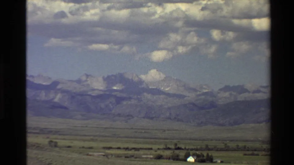 Western U.S. 1980 8mm wide view of a valley, small farm buildings, trees, and mountains beneath a cloudy sky.