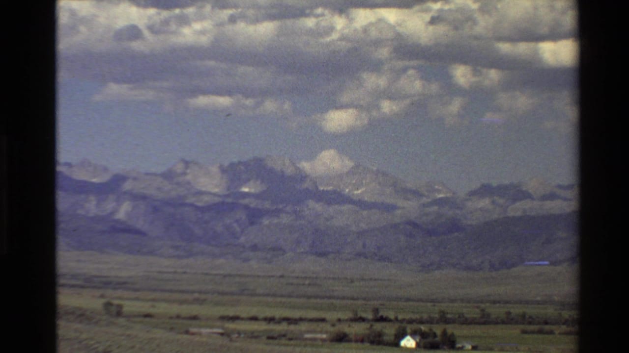1980 Western USA 8mm frame showing a wide plain, distant white building, foothills, and snow-streaked mountains under scattered clouds.