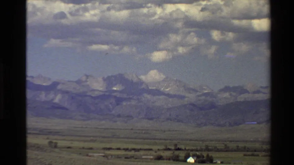 1980 Western USA 8mm frame showing a wide plain, distant white building, foothills, and snow-streaked mountains under scattered clouds.