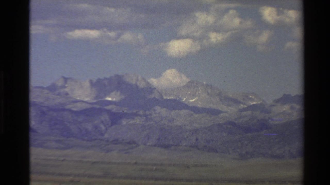 Mountain range and foothills under a partly cloudy sky, framed by dark film borders.