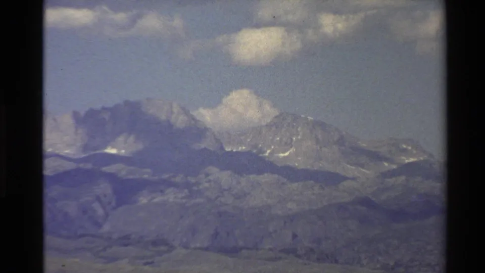 1980 Western USA 8mm frame showing a tighter zoom on gray mountain peaks with snow patches and a large cloud above.