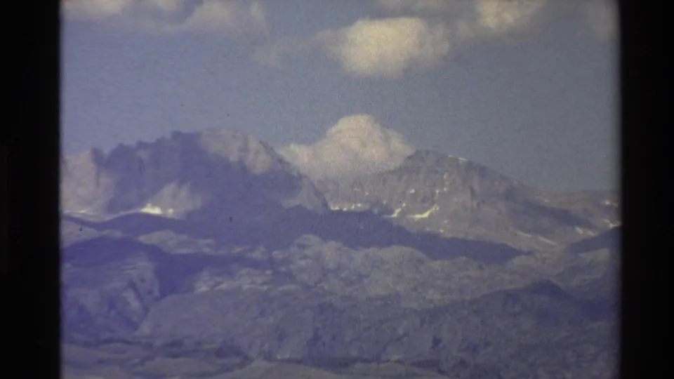 1980 Western USA 8mm frame at the end of the zoom, focused on snow-streaked peaks and layered foothills under a cloudy sky.