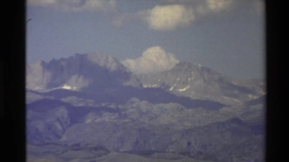Distant Western U.S. mountain ridge in a soft 8mm frame, with clouds over snow-streaked peaks and muted hills below.