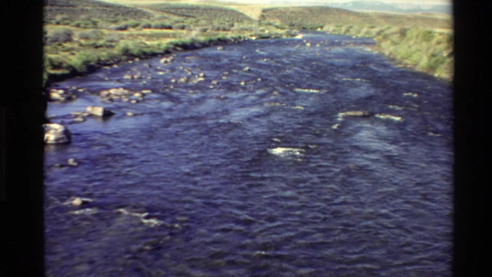 1980 8mm view of a flowing Western U.S. river with rocks, grassy banks, and low hills in the background.