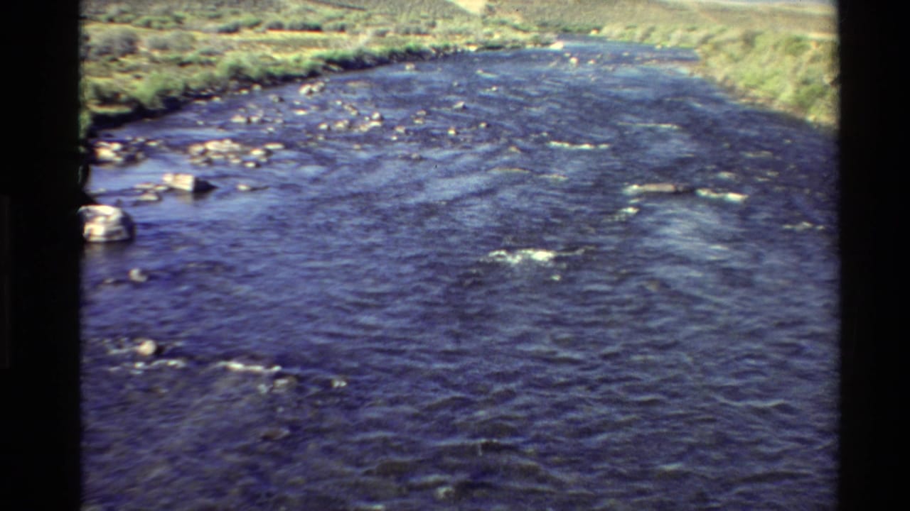 1980s 8mm view of a rocky Western USA river, with rippling water, pale stones, grassy banks, and dark film-frame edges.