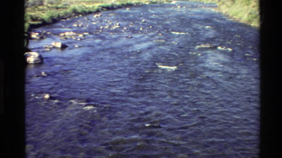 1980s 8mm frame of a river bend or channel in the Western USA, showing blue-purple water, scattered rocks, and grassy shoreline.